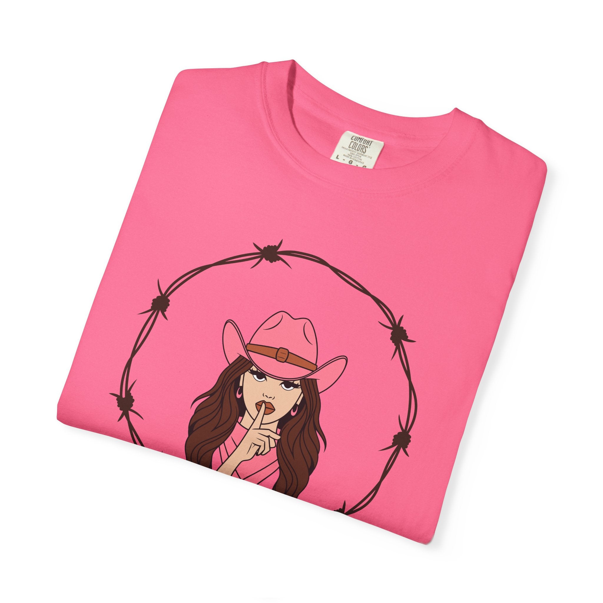 Folded Pink t-shirt with illustrated woman in pink cowboy hat and top making a “shh” gesture, framed by a circular barbed wire border; bold Western graphic tee with edgy feminine flair.