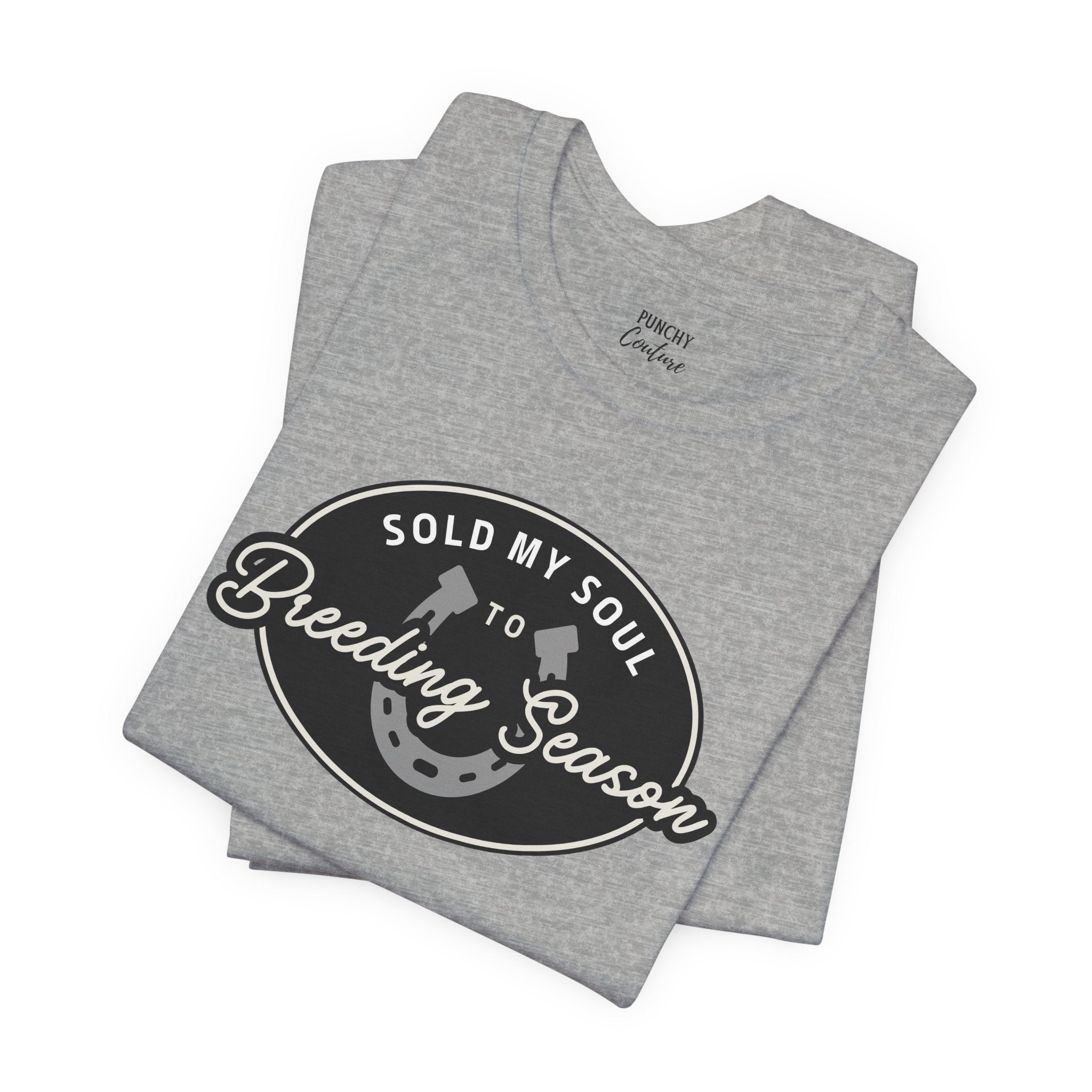 Folded gray t-shirt with black oval emblem featuring white and gray text “SOLD MY SOUL TO Breeding Season” and graphics of shovels and a horseshoe; humorous Western design with bold contrast.