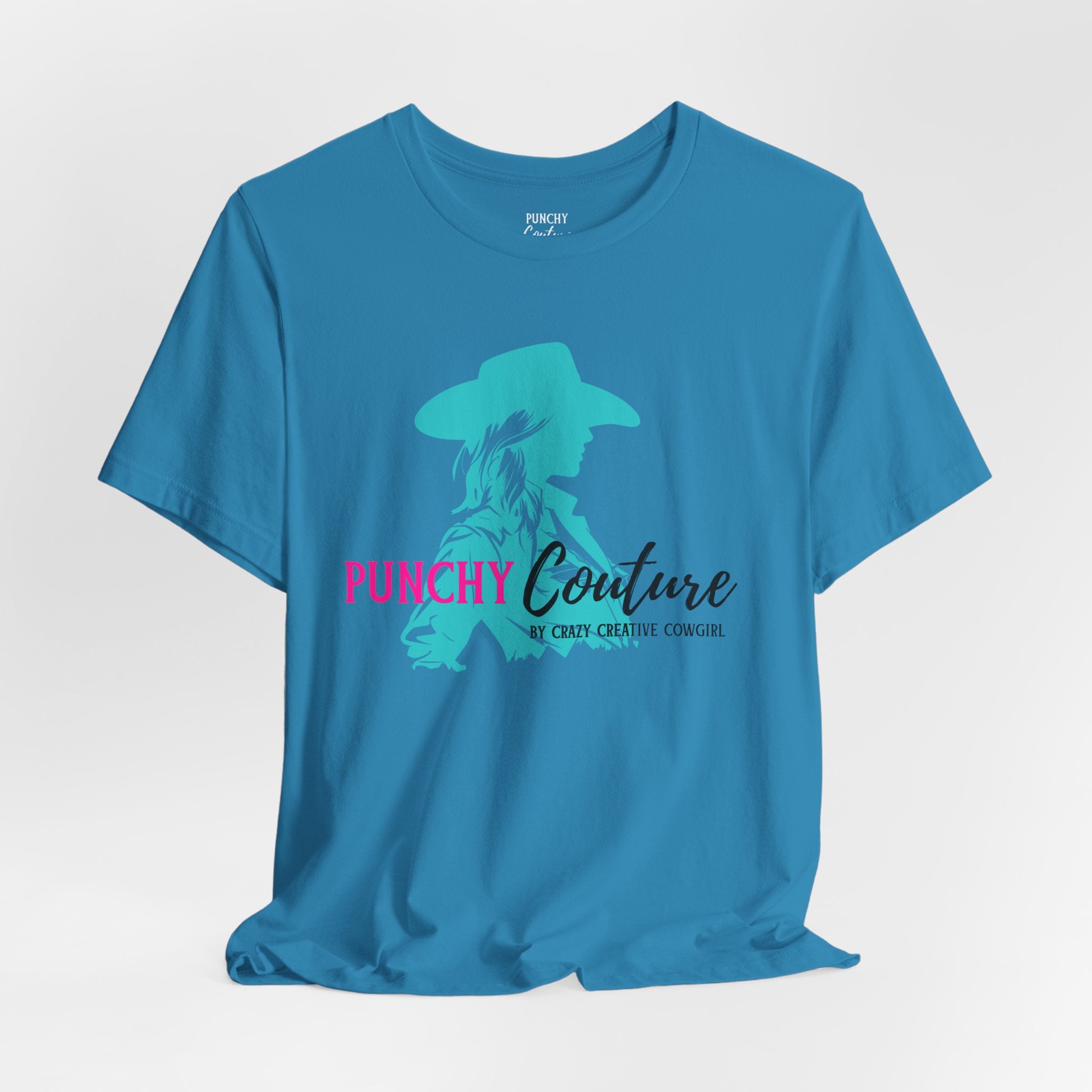 Blue t-shirt with turquoise cowgirl silhouette and bold pink "PUNCHY" text; features “Couture by Crazy Creative Cowgirl” tagline in black script and uppercase font; Western graphic tee with mythic brand energy.