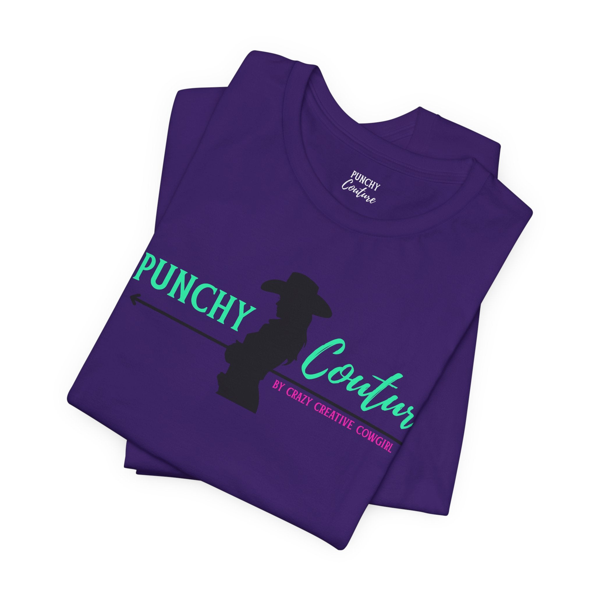 Folded purple t-shirt with silhouette of cowgirl holding a rope and bold green and turquoise “PUNCHY Couture” text; pink tagline reads “BY CRAZY CREATIVE COWGIRL”; Western graphic tee with mythic branding and bold feminine energy.