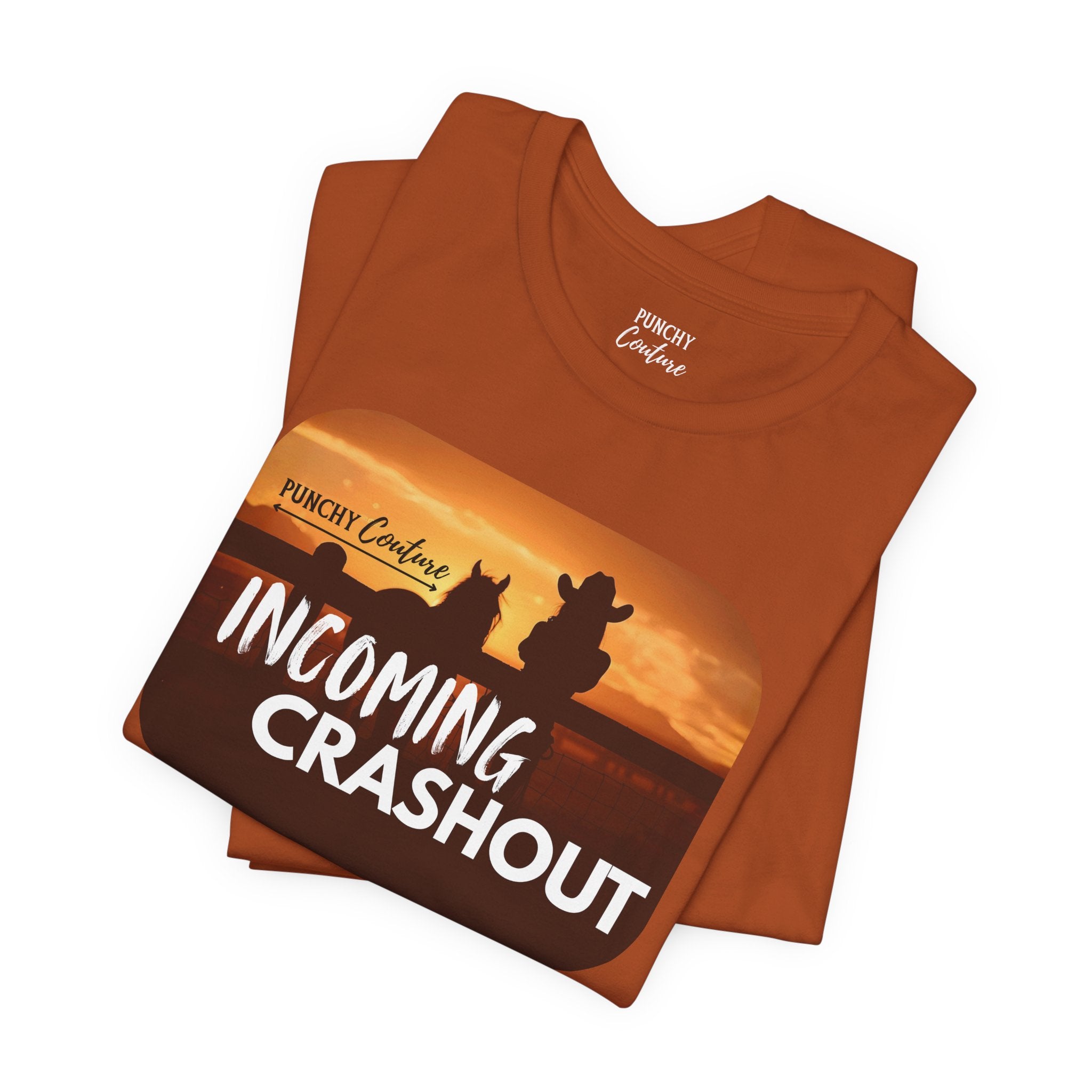 Rust-colored t-shirt with black silhouette of a cowboy and horse against a sunset, featuring stylized "PUNCHY Couture" text above and bold white "INCOMING CRASHOUT" below; Western graphic tee with dramatic flair and mythic energy.