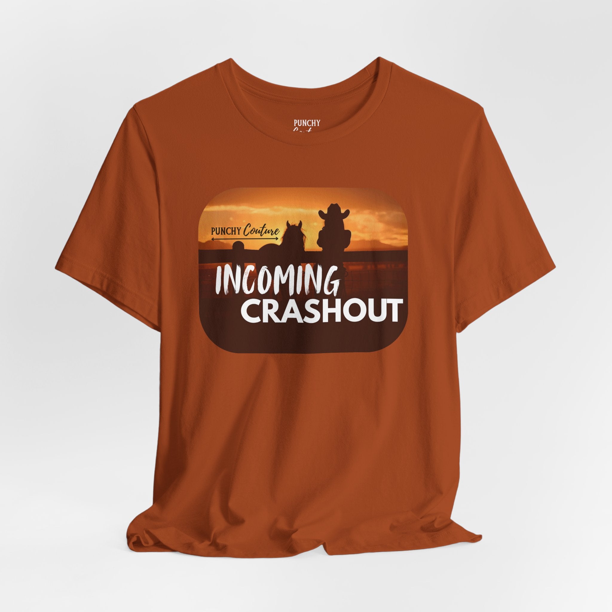 Rust-colored t-shirt with black silhouette of a cowboy and horse against a sunset, featuring stylized "PUNCHY Couture" text above and bold white "INCOMING CRASHOUT" below; Western graphic tee with dramatic flair and mythic energy.