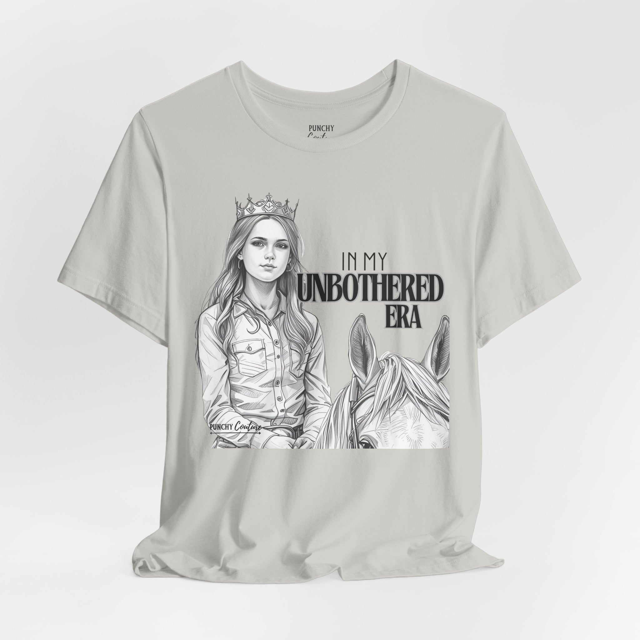 Light-colored t-shirt featuring a black-and-white illustration of a crowned cowgirl beside a horse, with bold text reading "IN MY UNBOTHERED ERA" and "PUNCHY COUTURE" below; a confident Western graphic tee celebrating sovereignty and style.