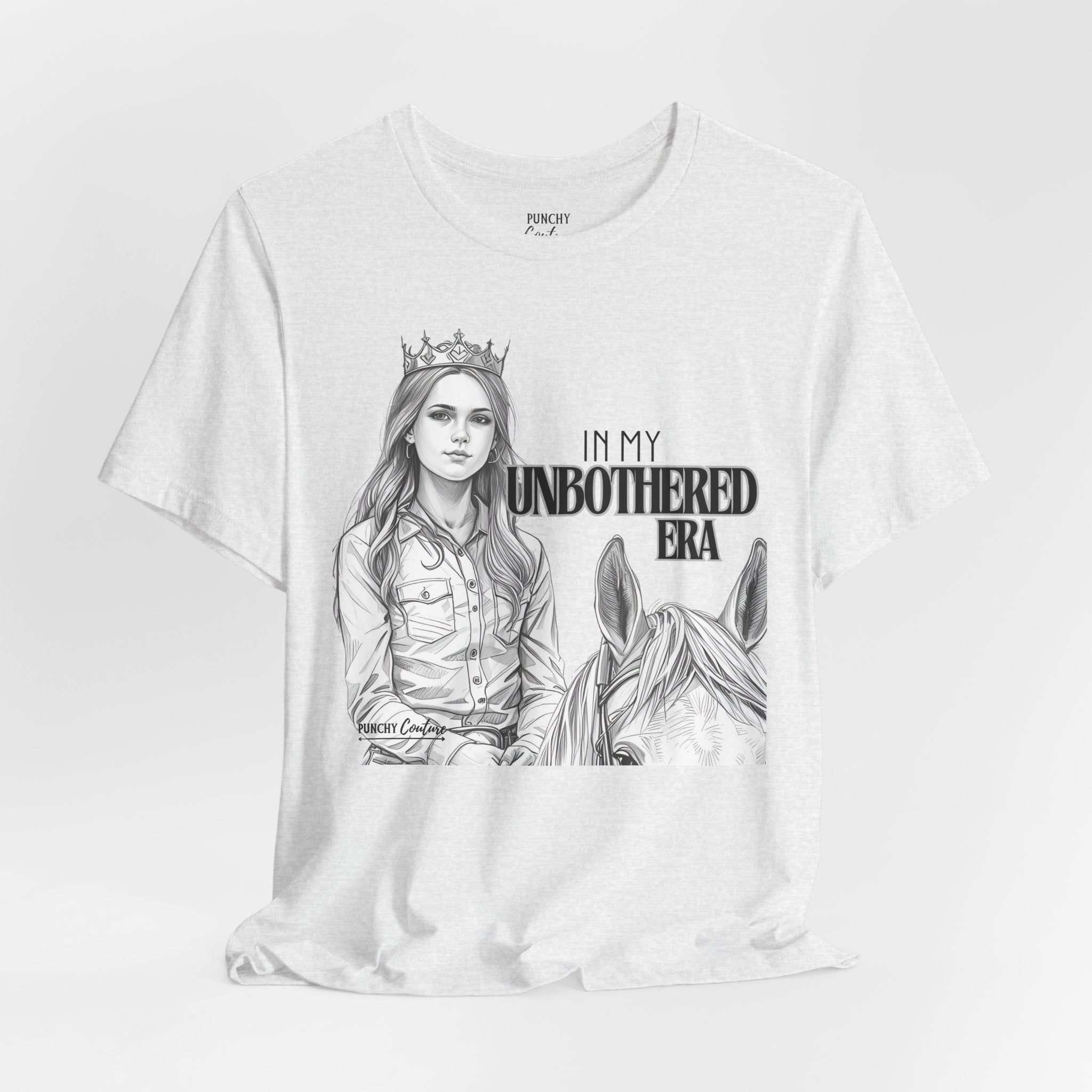 Light-colored t-shirt featuring a black-and-white illustration of a crowned cowgirl beside a horse, with bold text reading "IN MY UNBOTHERED ERA" and "PUNCHY COUTURE" below; a confident Western graphic tee celebrating sovereignty and style.